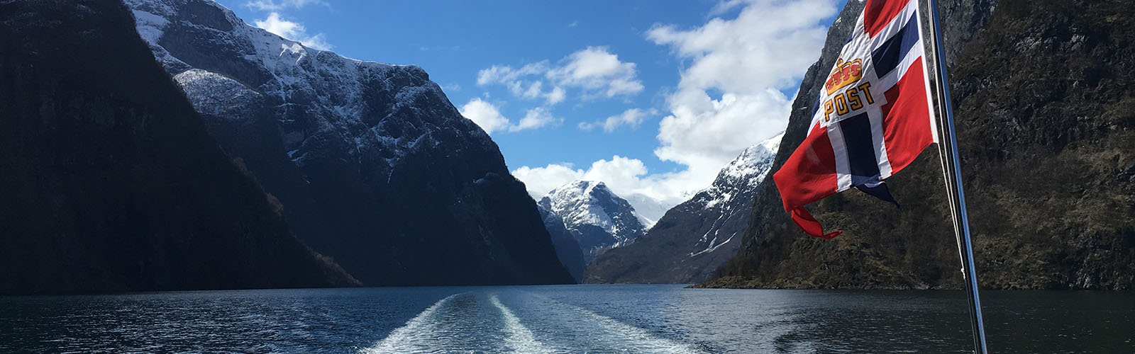 A Norwegian flag is visible from the back of a boat, which is created a wake in the water, and several mountains are visible in the background. 